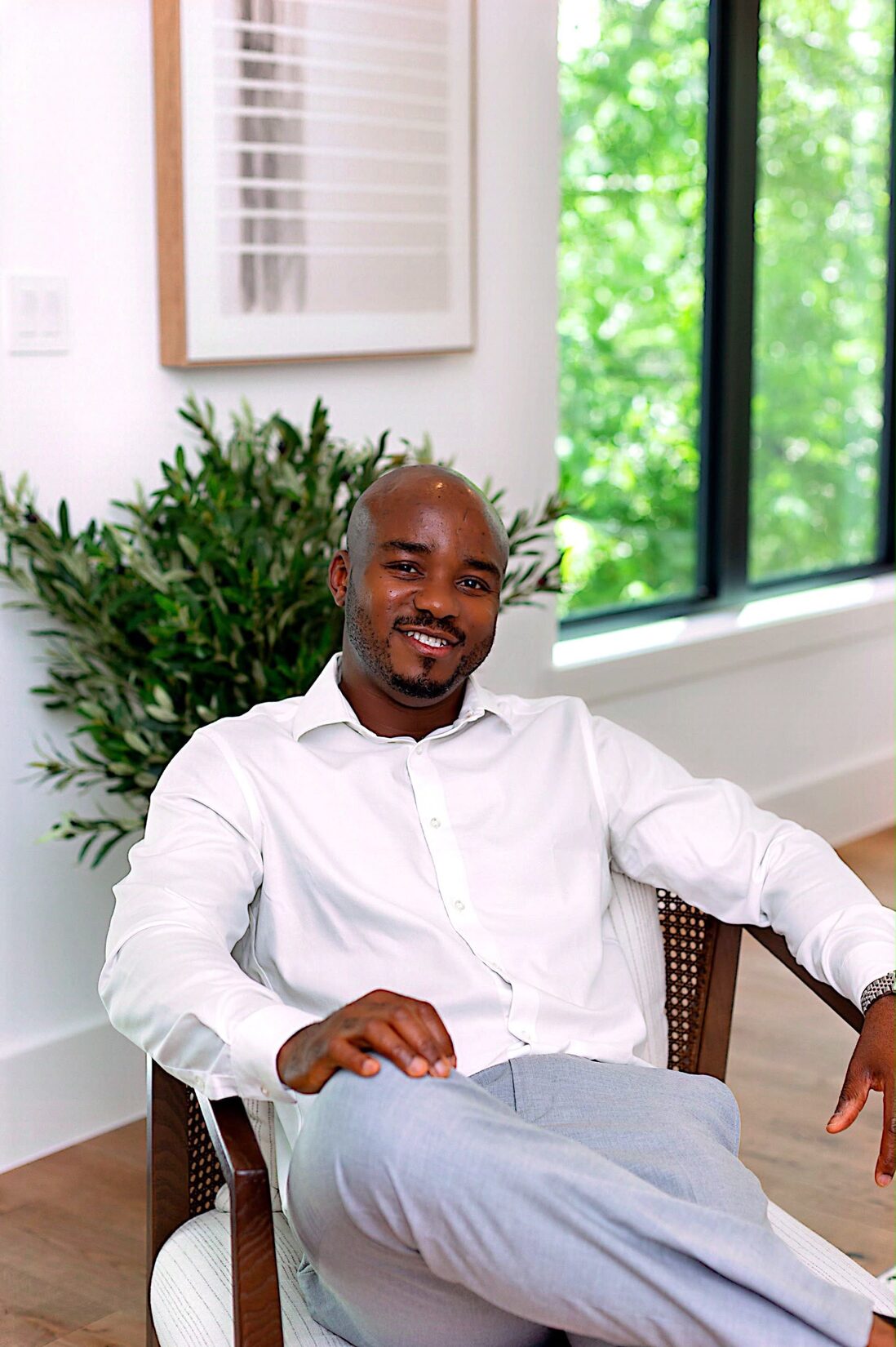 Henry S. Anaege smiling, seated in a cane chair by a window in a bright living room